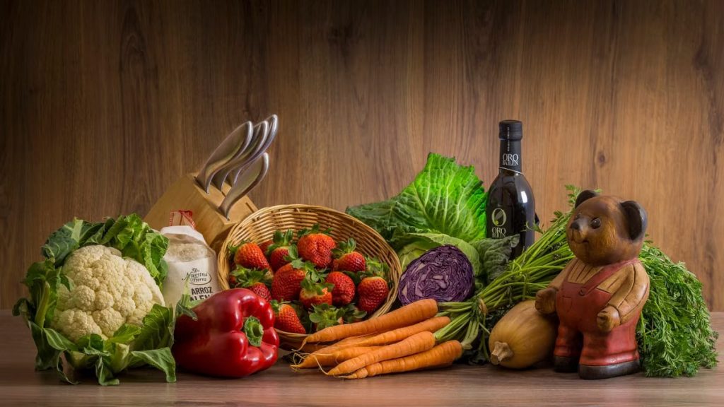 Arrangement of fresh vegetables and fruits on a wooden table with kitchen essentials.